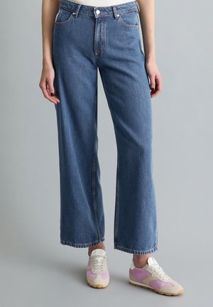 Niebieskie dżinsy o prostych nogawkach noszone z białymi i różowymi siateczkowymi sportowymi butami na osobie stojącej na jednolitym szarym tle.