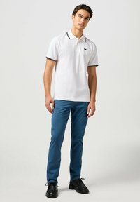 Weißes Polohemd mit schwarzem Trim, kurzen Ärmeln und gesticktem Logo; kombiniert mit blauen Hosen und schwarzen Schnürschuhen.
