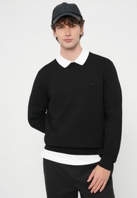 Czarny sweter dziany z białą koszulą z kołnierzykiem pod spodem. Sweter ma długie rękawy i luźny krój. Na piersi znajduje się subtelne logo marki.