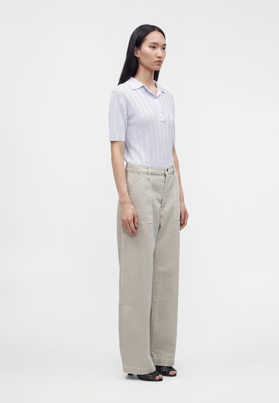 Femme debout portant un polo à manches courtes rayé clair, un pantalon large beige et des chaussures noires à bouts ouverts sur fond blanc.