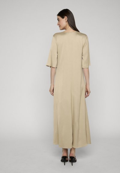Femme aux longs cheveux noirs debout, tournée de dos, portant une robe midi beige à manches mi-longues et des sandales à talons noires.