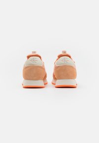 Jasno-pomarańczowe zamszowe sneakersy z kremowym akcentem. Posiadają wyściełany kołnierz, wytłoczone logo na pięcie oraz białą podeszwę środkową z pomarańczową podeszwą zewnętrzną.