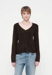Mladostnik s kratkimi rjavimi lasmi, oblečen v temno V-izrezano pulover in svetlo modre jeans hlače, ki stoji ob belem ozadju.