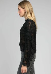 Blouse noire transparente avec des motifs floraux texturés, des accents froncés, un col montant et des manches longues, associée à un jean gris taille haute.