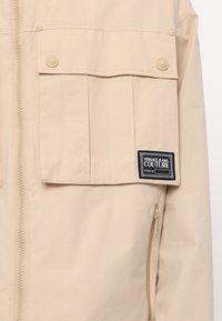 Beige outerwear jacket made of smooth fabric, featuring a front zipper, two large flap pockets, and a branded label patch.