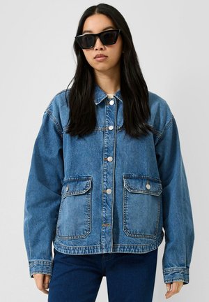 Frau mit langen dunklen Haaren, die große schwarze Sonnenbrillen, eine übergroße blaue Jeansjacke mit Fronttaschen und dunkelblaue Jeans trägt.