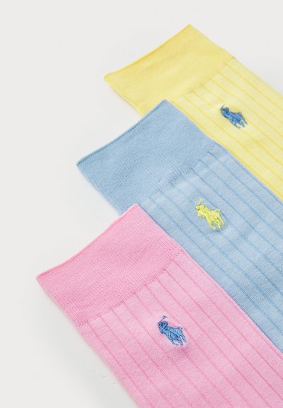 Trois paires de chaussettes côtelées en rose, bleu et jaune. Chacune présente un petit logo brodé et une texture douce et lisse.