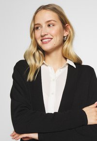 Melnā džemperī ar garām piedurknēm virs baltas pogājamas krekla. Džemperis ir gludas tekstūras un brīvā piegriezumā, ar vienkāršu dizainu.