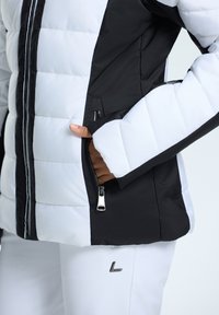 Zwart-witte puffer jacket met horizontaal gestikt ontwerp, zijzak met rits en een gladde textuur. Witte broek met metalen accenten.