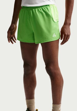 Man draagt felgroene sportshorts met een klein logo, beige sokken en een witte cropped shirt boven de taille, staand tegen een effen achtergrond.
