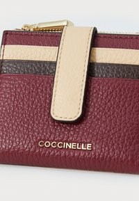Leather cardholder in burgundy, beige, and dark brown with a textured finish. Features a gold zipper and an embossed logo at the front.