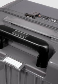 Valigia rigida grigia con manico nero, due cerniere e porta-etichetta per il nome. Presenta una superficie texturizzata e una serratura a combinazione integrata.