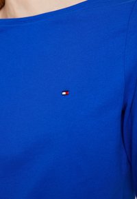 Blå bomullsskjorta med rund halsringning och korta ärmar, prydd med en liten broderad logotyp i röd, vit och blå på bröstet.