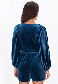Mélykék bársony overál puffos hosszú ujjakkal, fodros vállakkal és rugalmas derékrésszel. Texturált anyagból készült, rövid szárú alsó résszel.