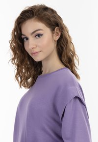 Camicia leggera a maniche lunghe di colore viola con scollatura rotonda e polsini arrotolati, caratterizzata da una texture liscia e morbida.