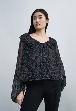 Mladá žena nosi čiernu priehľadnú blúzku s bielymi bodkami a volánikovým výstrihom, v kombinácii s priliehavými čiernymi nohavicami na jednoduchom pozadí.