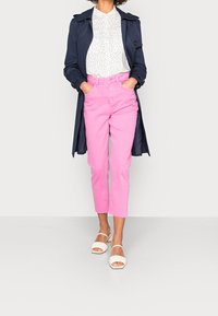 Abrigo de trench marino, blusa blanca con lunares, pantalones cortos rosas y sandalias blancas con acentos rizados. Presenta un corte recto y dobladillos deshilachados.