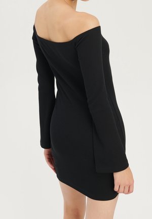 Femme portant une robe noire moulante, épaules dénudées, manches longues, vue de dos sur un fond uni.