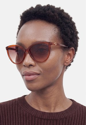 Personne aux cheveux courts et bouclés portant de grandes lunettes de soleil teintées marron et un pull côtelé marron foncé sur un fond uni.