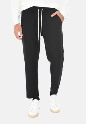 Uomo con pantaloni neri con coulisse, mani nelle tasche, sneakers bianche e una maglietta bianca a maniche lunghe su uno sfondo bianco.