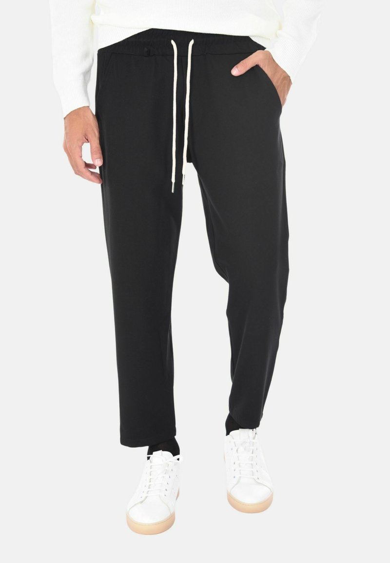 Uomo con pantaloni neri con coulisse, mani nelle tasche, sneakers bianche e una maglietta bianca a maniche lunghe su uno sfondo bianco.