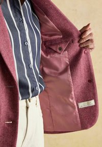 Osoba v bielych nohaviciach a modrej pruhovanej košeli otvorila ružový tweedový kabát, ktorý ukazuje jeho saténovú podšívku s vnútornou gombíkovou kapsou.
