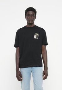 Camiseta negra de cuello redondo y mangas cortas. Presenta un logo impreso en el lado izquierdo del pecho. La tela parece suave y casual.