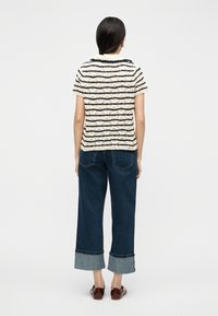 Γυναίκα με μακριά σκούρα μαλλιά, φορώντας ένα μπεζ και μαύρο ριγέ πουλόβερ με κοντά μανίκια, φαρδιά μανσέτα σε σκούρο μπλε τζιν και καφέ παπούτσια, στέκεται με το πρόσωπο προς τα πίσω.