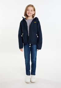 Marinblå zip-up hoodie med texturerad finish, vit foder och dragsko-accenter. Bärs över en grå skjorta och blå jeans, tillsammans med vita skor.