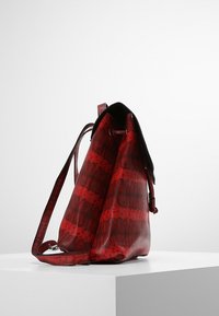 Punainen käärmeenluomukuvioinen laukku, jossa on teksturoitu pinta, nyörikiristys ja pitkä säädettävä olkahihna. Pyöristetty muoto helpottaa pääsyä laukkuun.