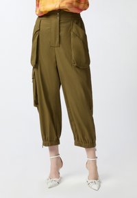 Maslinaste cargo hlače s visokim strukom, prevelikim džepovima, elastičnim manžetama i prednjim zatvaranjem na dugme. Uparene s bijelim sandalama na visoku petu.