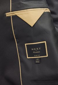 Doublure de costume noire à motif avec bordure beige, avec une étiquette mentionnant "NEXT Occasion Slim Fit", taille 40R, fabriquée au Vietnam.