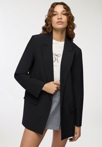 Blazer nero con rever a punta, chiusura a un bottone, due tasche anteriori e vestibilità rilassata. Indossato su una t-shirt bianca con grafiche.