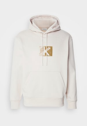 Light beige hoodie made of soft fabric, featuring a front pocket and drawstring hood. Centered gold logo on chest in a square design.