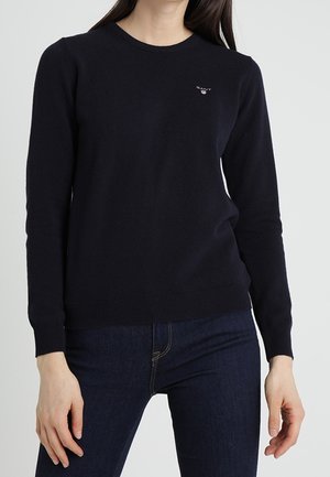 Femme portant un pull noir ajusté à manches longues et un jean bleu foncé, debout devant un fond blanc uni.