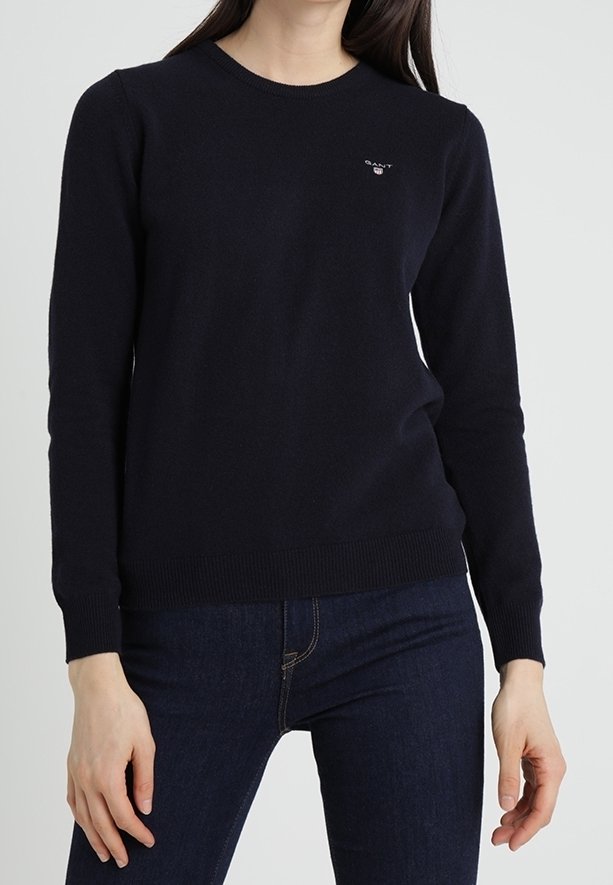 Femme portant un pull noir ajusté à manches longues et un jean bleu foncé, debout devant un fond blanc uni.