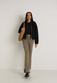 Crna skraćena jakna, rebraste maslinasto zelene hlače i crne cipele. Smeđa torba s zakrivljenim dizajnom. Jednostavna pozadina, model hoda unaprijed.