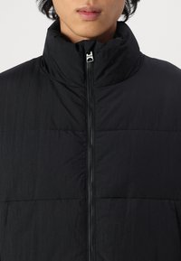 Black puffer jacket with a high collar, featuring a full zipper and textured fabric, showing a quilted design and minimal hardware details.