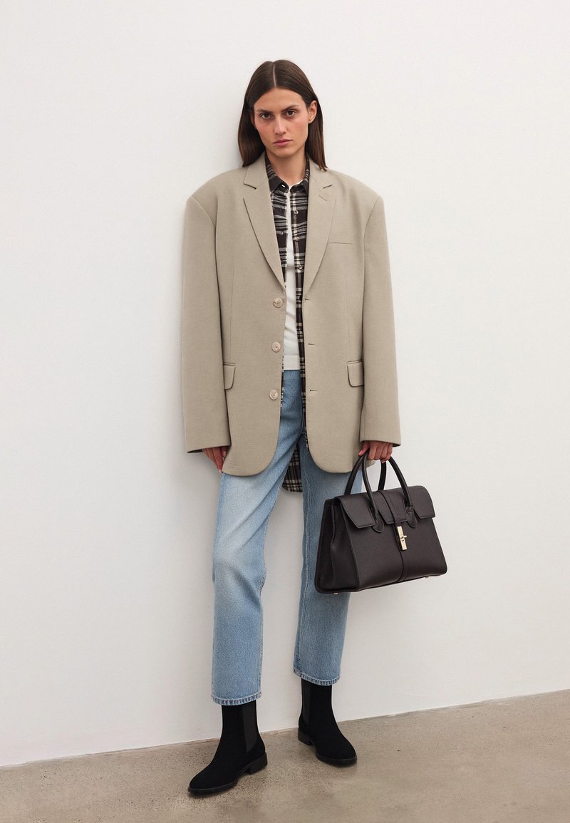 Mujer con blazer beige oversized, camisa de cuadros, jeans de mezclilla azul claro, botas negras, sosteniendo un bolso de cuero negro, de pie frente a una pared blanca.