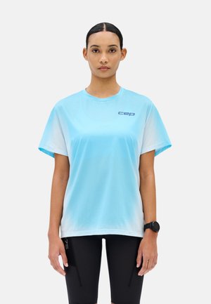 Vrouw draagt lichtblauw sportshirt met korte mouwen en "cep"-logo, en zwarte fietsshorts, staand tegen een effen lichte achtergrond.