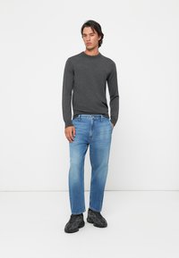 Chemise gris à manches longues, jeans bleu droit et baskets noires portés par un mannequin masculin en position debout, fond neutre.