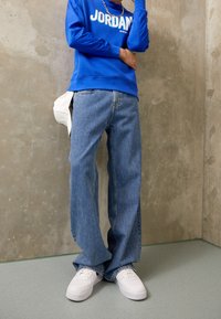 Blå sweatshirt med vit text "JORDAN", kombinerad med ljusblå denimjeans och vita sneakers, står mot en texturerad betongvägg.