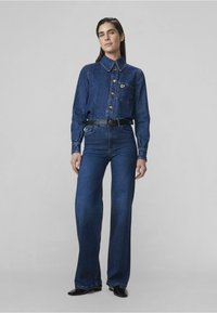 Denim-paita kauluksella, nappisuljenta ja rintatasku. Korkeavyötäröiset siniset farkut suoranlahkeena, musta vyö ja tummat kengät.