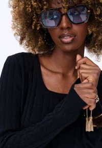 Czarny sweter z prążkowanej dzianiny z szerokim dekoltem. Przesadzone kwadratowe okulary przeciwsłoneczne z przyciemnianymi szkłami. Kręcone włosy i złączone ręce, prezentujące pierścionek.