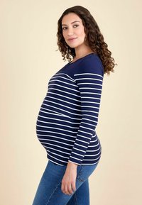 Tumed ja valged triibulised pikkade varrukatega rasedustops, mis on vöökoha juurest kitsas, kangas on sile, kombineeritud siniste denimiste teksadega.