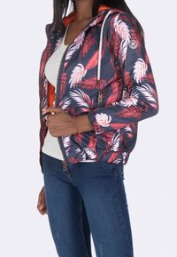Floral-patterned jacket in navy and red, featuring a zip front, adjustable hood with drawstrings, and elastic cuffs. Worn over a light top.