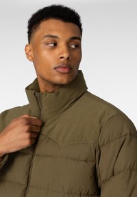 Olivenfarget polstret jakke med høy krage, med ribbede detaljer på skuldrene og fronten, samt glidelås. Glatt tekstur, lettvektig.