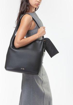 Donna in abito grigio senza maniche che tiene una grande borsa nera in pelle con una pochette in rilievo attaccata sulla spalla.