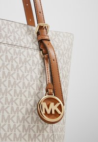 Sac cabas beige avec impression de logo, bandoulière en cuir marron, ferrures dorées et pendentif circulaire MK. Tissu texturé avec motif détaillé.