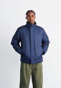 Navy blue padded jacket with high collar, featuring a front zipper, two side pockets, and a smooth texture. Worn over olive green pants.